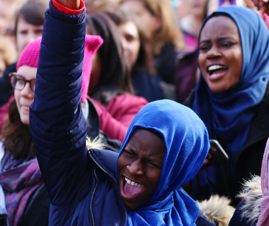 A Muslim woman in a blue hijab raises her fist in the air at a rally
