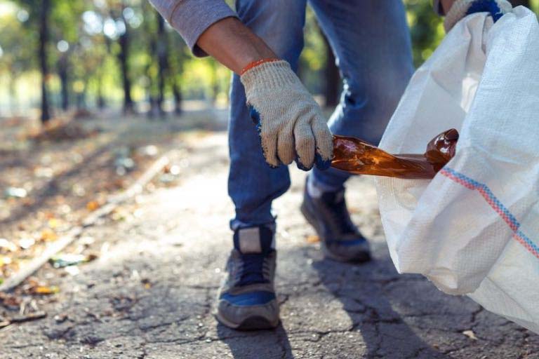 A man picks up a glass bottle and puts it in a white garbage bag in a park