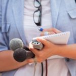 A journalist holds a notebook, a recorder, and two microphones