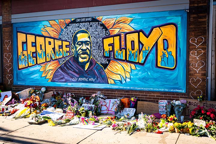 A brick wall mural with "George Floyd" in large block letters and an image of George Floyd. Flowers, notes, and posters were placed on the sidewalk below.