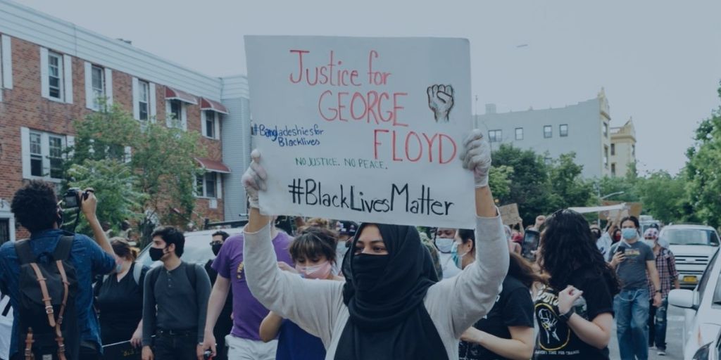 A Muslim woman wearing a mask in a crowd of protesters holding a sign that reads "Justice for George Floyd #BlackLivesMatter"