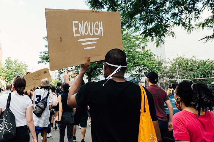 The back of a masked Black Lives Matter protester holding up a cardboard sign that reads "ENOUGH"