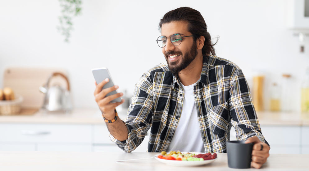 A smiling man browses his smartphone while enjoying a meal and mug of coffee while sitting in a bright kitchen