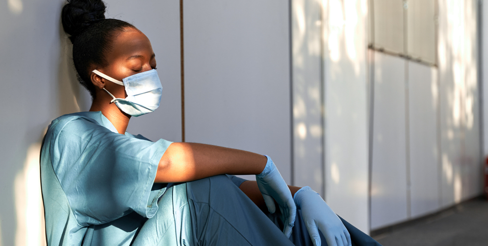 Tired female healthcare professional in a mask and blue scrubs sits against a wall with her eyes closed