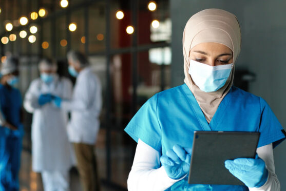 A masked female healthcare worker wearing a hijab, blue scrubs, and medical gloves works on a digital tablet while three colleagues meet in the background