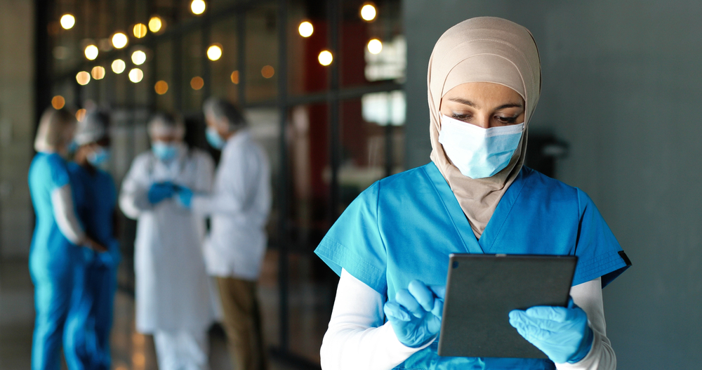 A masked female healthcare worker wearing a hijab, blue scrubs, and medical gloves works on a digital tablet while three colleagues meet in the background