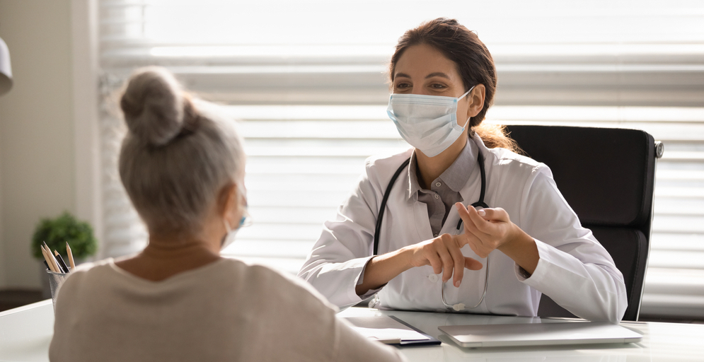 Female doctor at her desk counsels a patient, both wearing masks