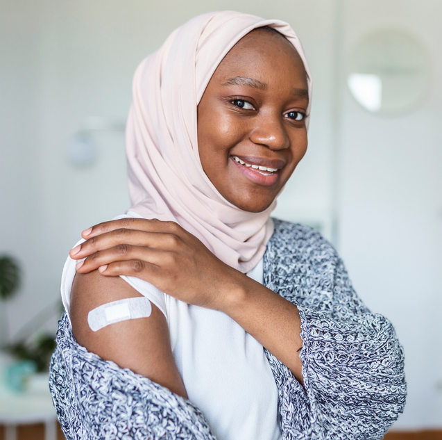 A woman wearing a pink hijab smiles showing a bandaid covering a vaccine or medicine injection site on her arm.