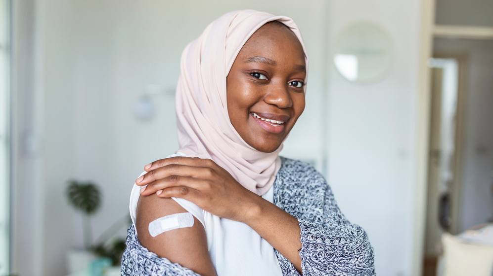 A woman wearing a pink hijab smiles showing a bandaid covering a vaccine or medicine injection site on her arm.