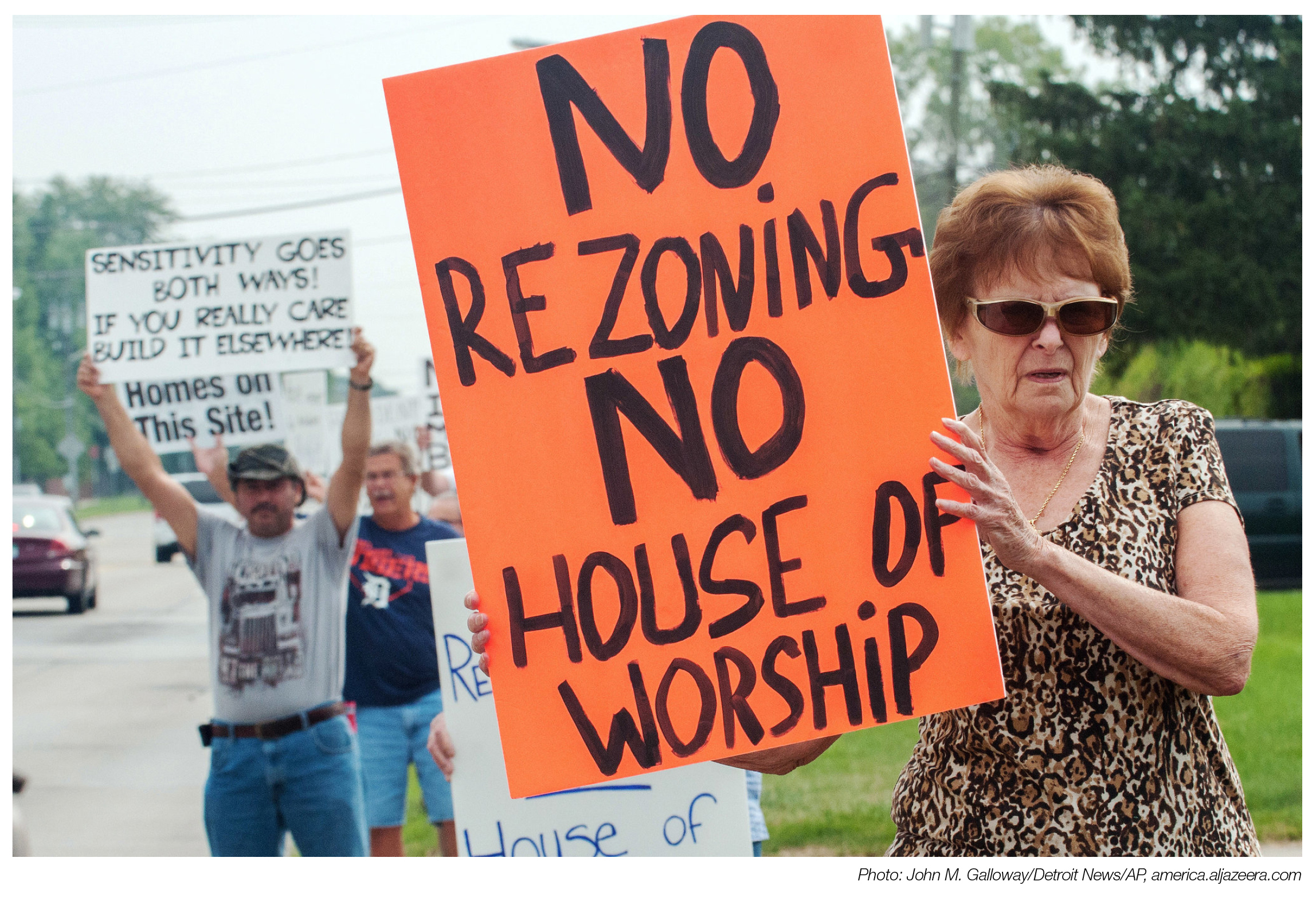A woman holds an orange sign reading "no rezoning, no house of worship" with several people behind her, also holding signs.