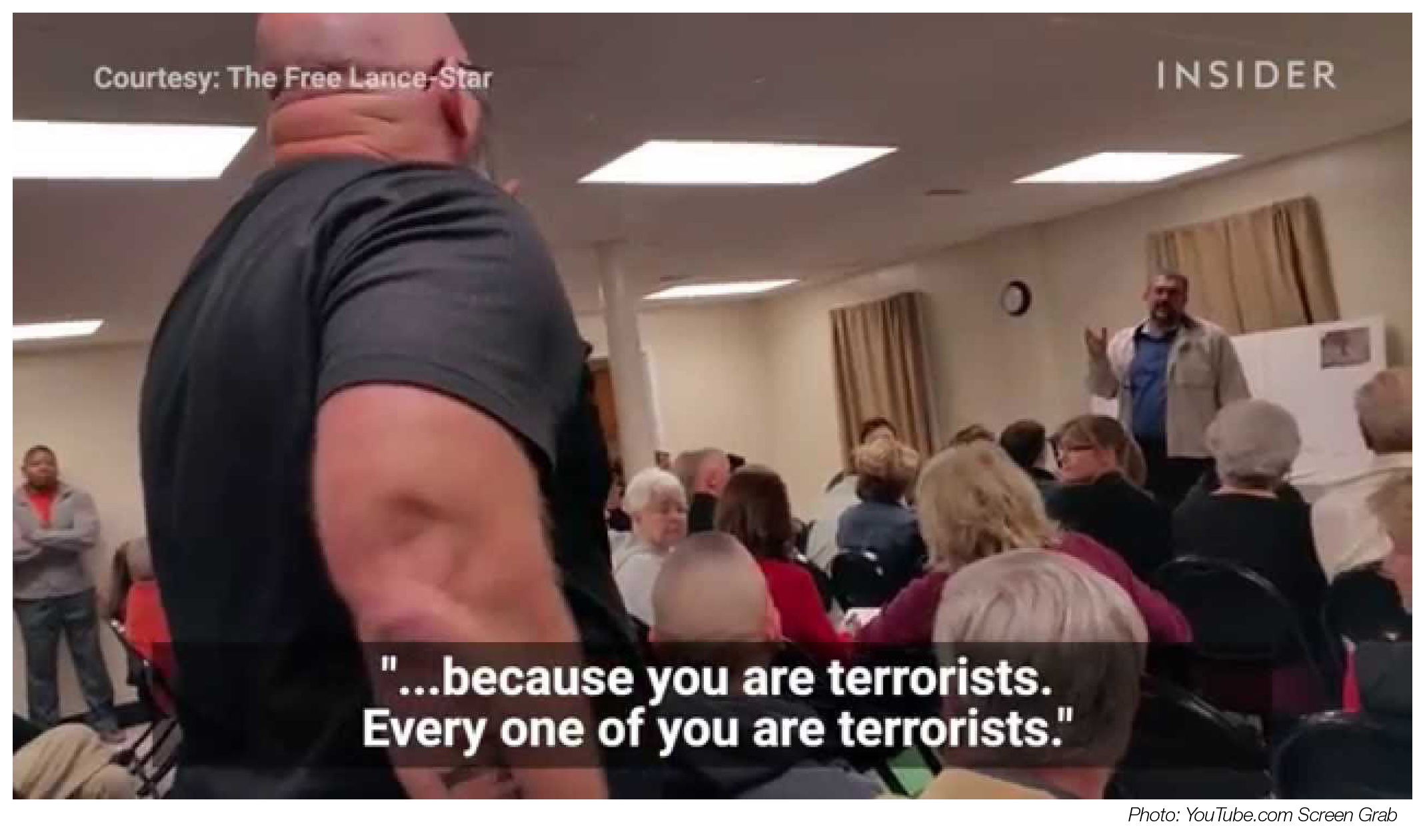 In a community gathering, a man stands and gestures at a speaker in the distance. Image caption reads: "because you are terrorists, every one of you are terrorists."