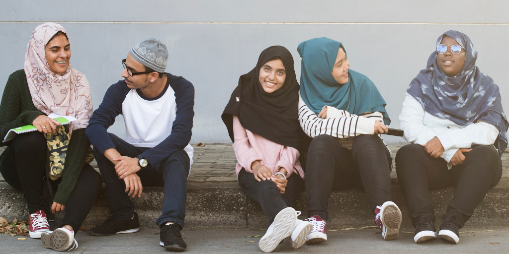 Five Muslim youth sit outside and talk against a wall