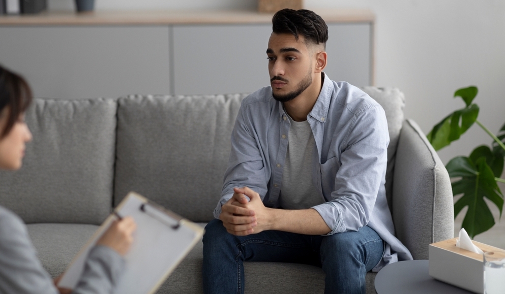A worried looking man sits on a couch for a consultation with a healthcare professional.