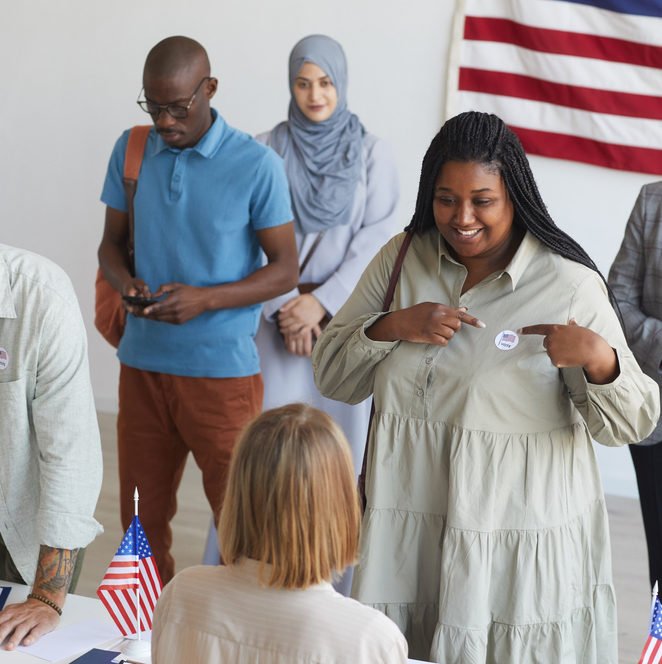 Group of people registering at polling station decorated with American flags on election day, focus on smiling woman pointing at I VOTE sticker.