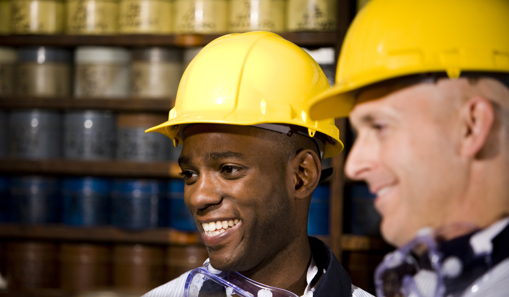 Two coworkers wearing yellow hardhats in a printing shop in front of shelves.