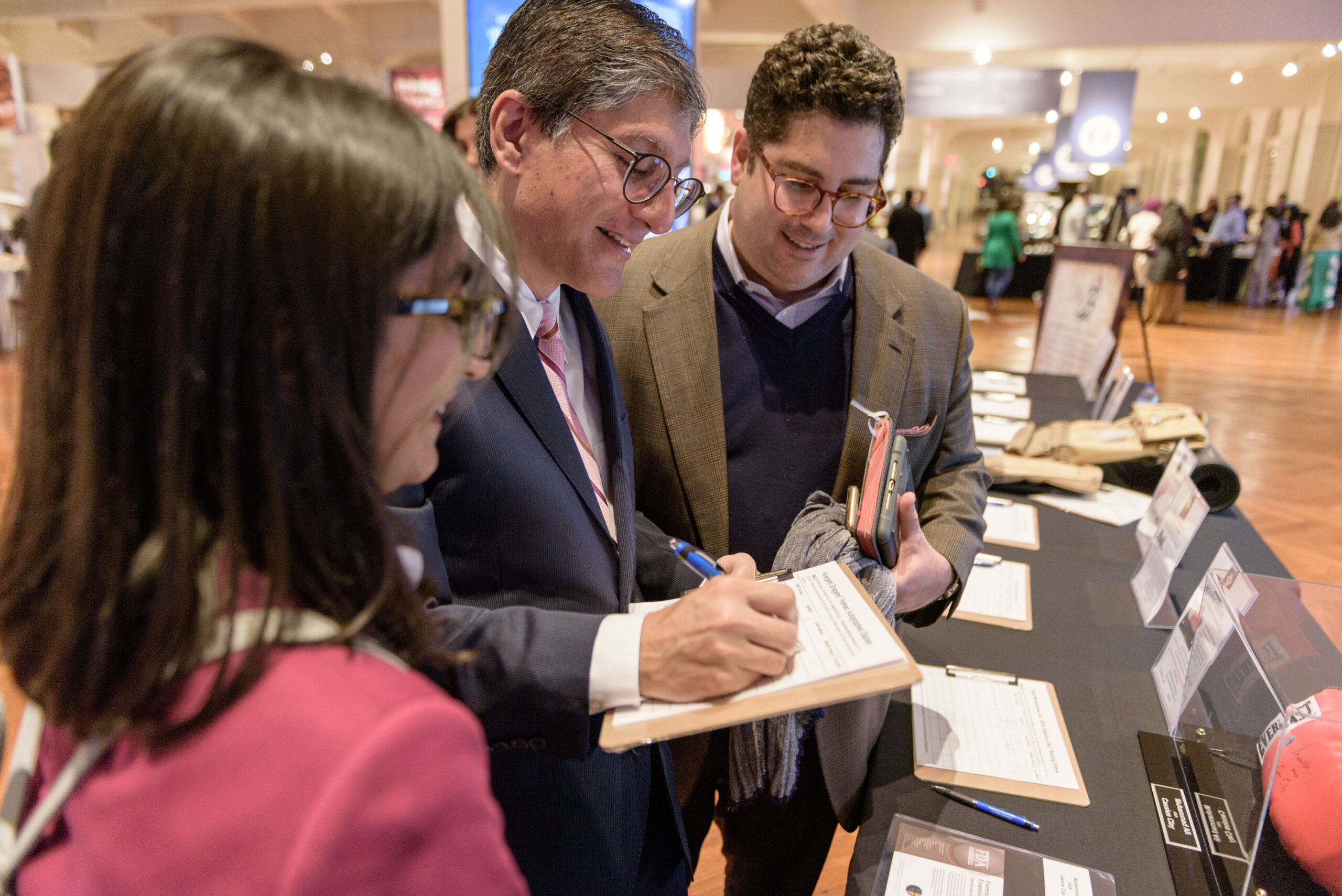 At a promotional table, two men and a woman stand closely, smiling, looking down at a paper on a clipboard.