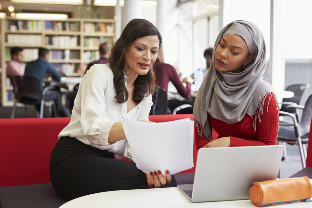 A female college student wearing a hijab reviews papers with a teacher.