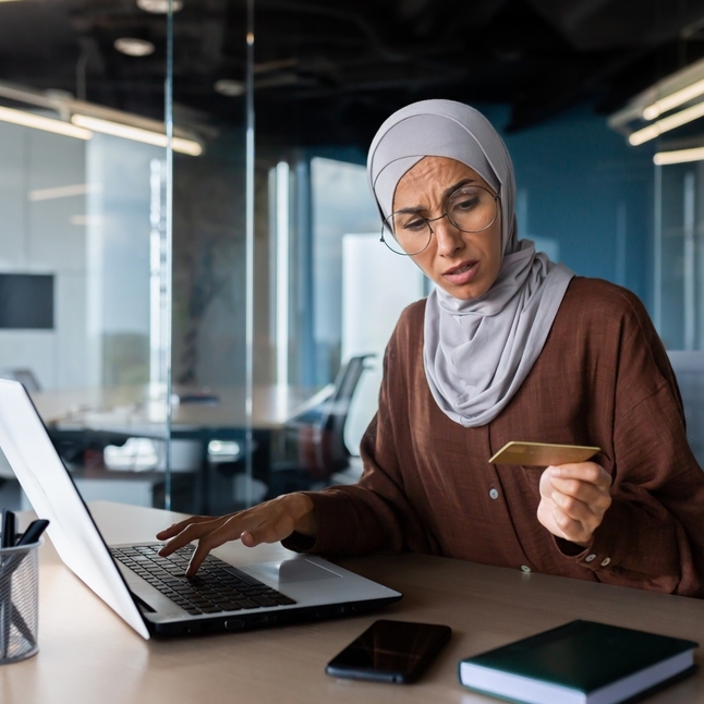 A woman in a hijab looks concerned while holding a credit card in her office.