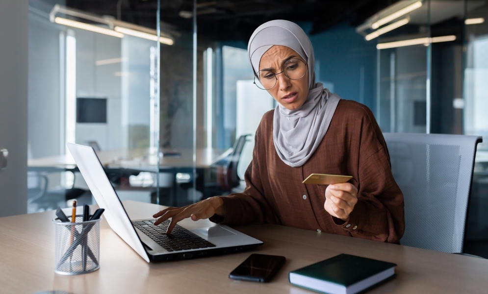 A woman in a hijab looks concerned while holding a credit card in her office.