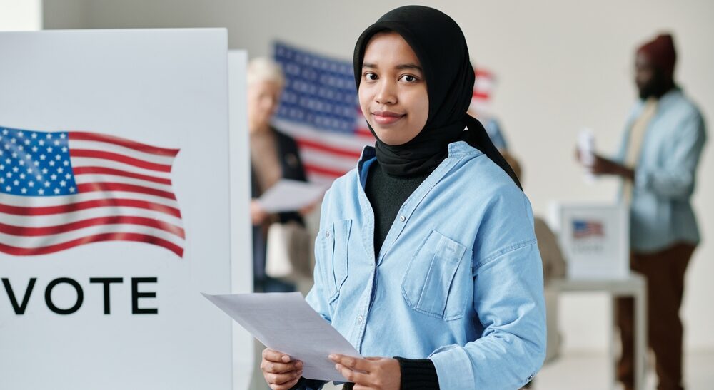 A young woman in a hijab and blue jacket holds voting papers next to a voting booth with the American flag.