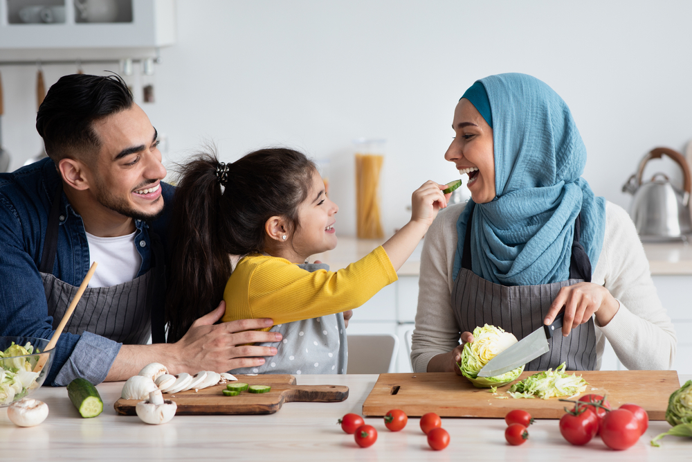 A young family of three prepares a meal together in the kitchen. They're all smiling as the couple's daughter feeds her mother a sliced cucumber.