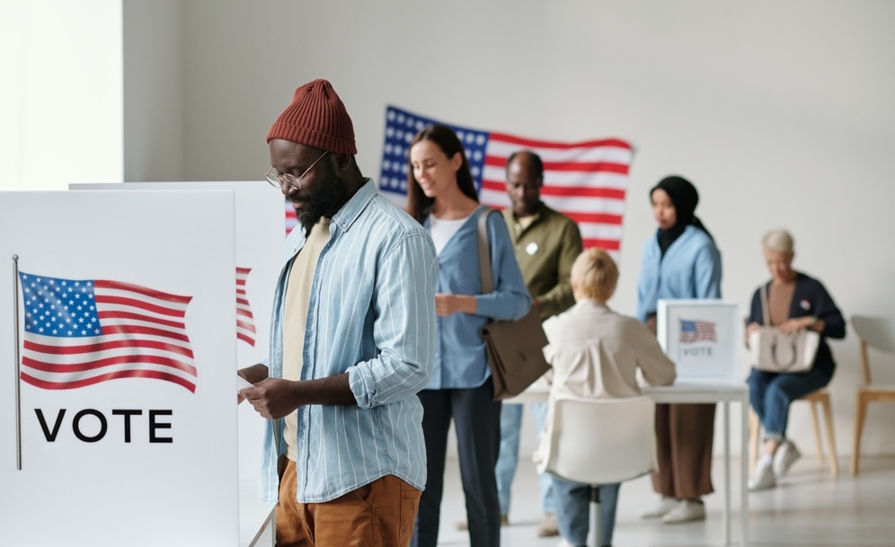 A group of young voters in casual clothing stands in a queue along voting booths and puts their ballots into boxes